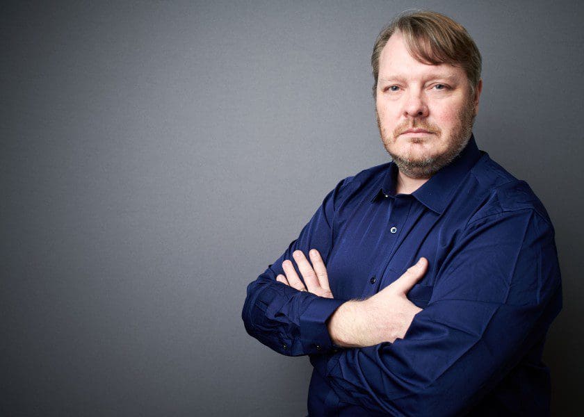 A professional photograph of Simon Marshall. He is facing the camera with his arms crossed, and is wearing a dark blue shirt.