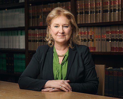 Dame Sue Carr sitting at a desk. She is wearing a green blouse and a black jacket. Behind her are shelves of law books.